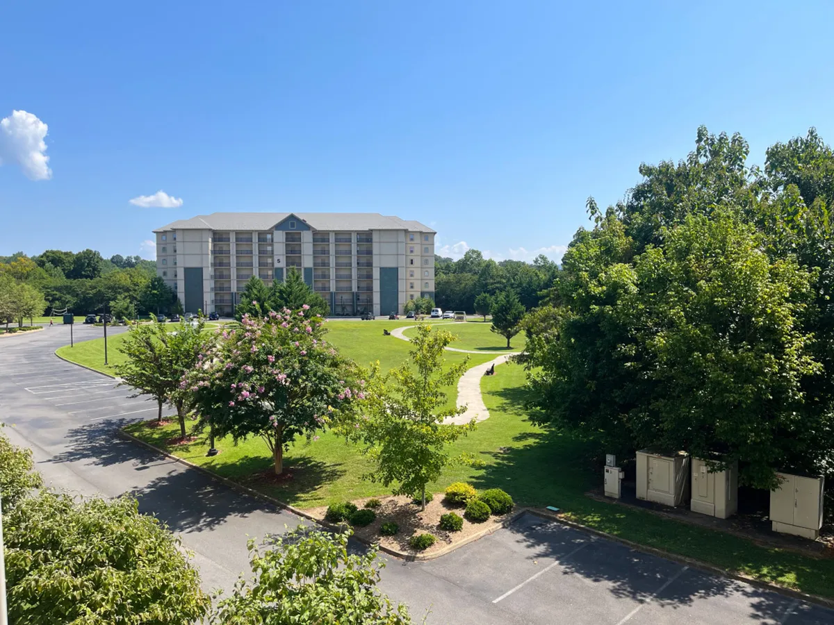 A multi-story building set against a backdrop of lush green trees and a well-maintained lawn, with a walking path leading across the grass. The scene is inviting, with ample space for walking, playing, or gathering outdoors.