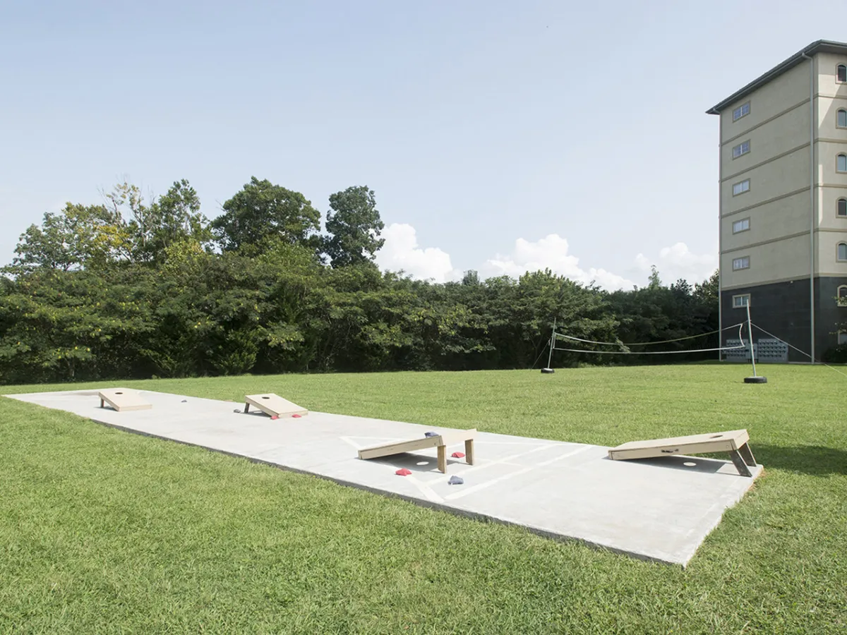 An expansive lawn with bean bag toss (corn hole) boards set up, shaded by mature trees and with the main building visible in the background. The scene invites friendly outdoor games and social connection in a natural setting.