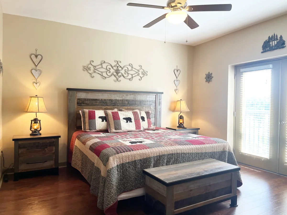 The image showcases a bright, inviting bedroom with a plush bed dressed in a patterned quilt, flanked by warm nightstands and softly glowing lamps. A large window with blinds allows natural light to fill the space, while decorative wall art adds a touch of mountain-inspired charm. The scene feels cozy and welcoming, perfect for relaxation and unwinding after a day of adventure.