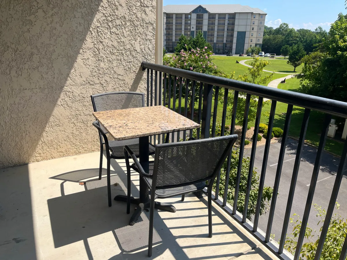 A peaceful private balcony scene with a granite-topped table and two metal chairs overlooking a landscaped area. The setting promises relaxation and a gentle connection to the outdoors, ideal for morning coffee or evening chats.