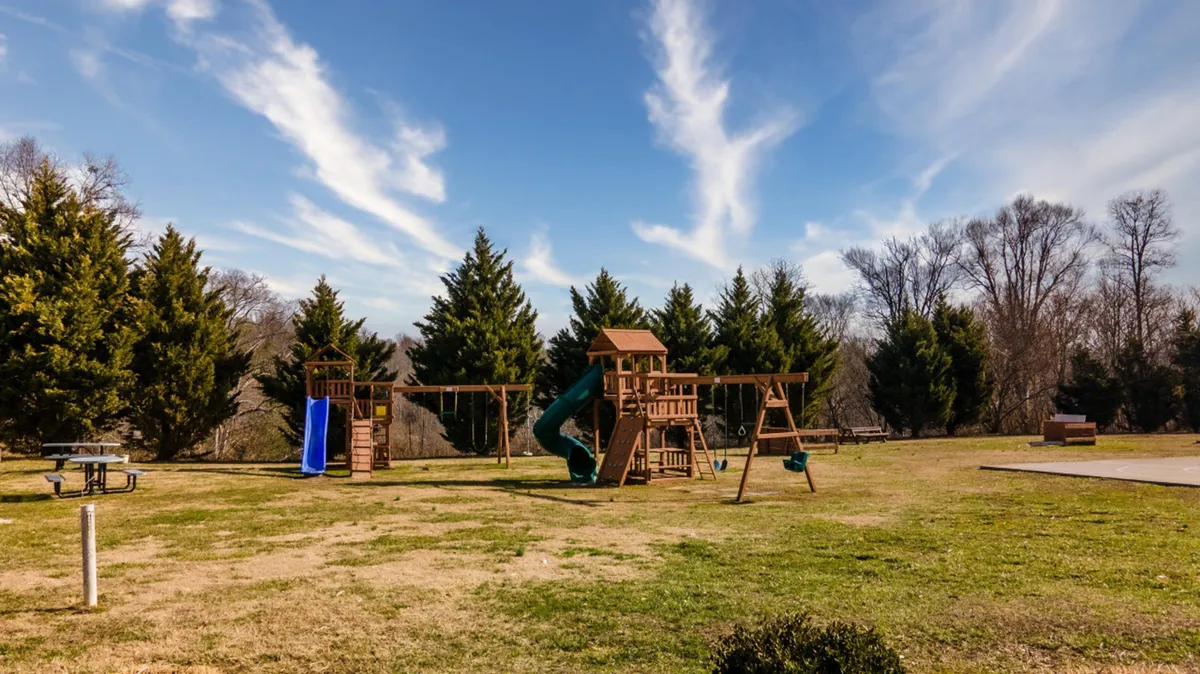 A vibrant playground set in a green, grassy area surrounded by trees. A picnic table nearby welcomes families for snacks and gatherings, while children play on the equipment, emphasizing outdoor fun and social interactions.