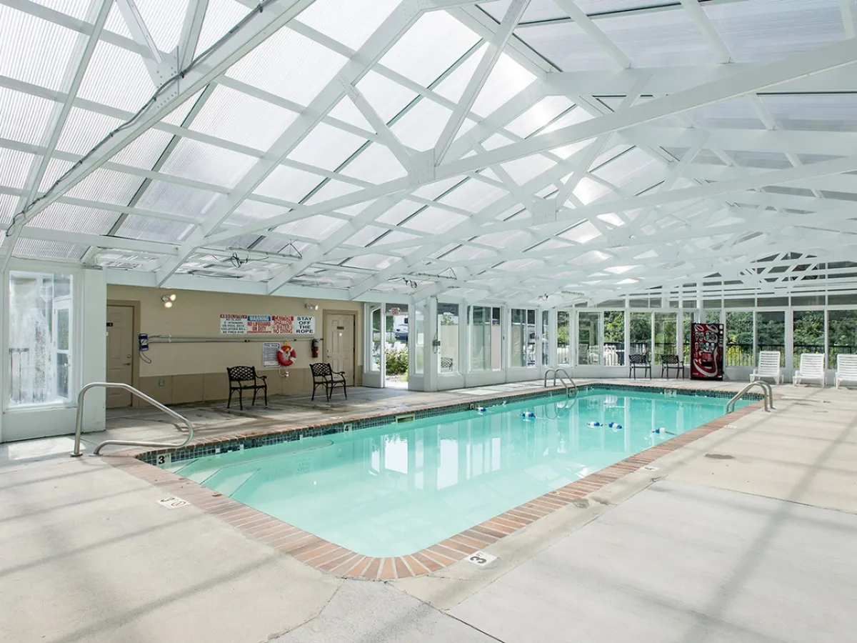 A bright indoor swimming pool area, covered by a transparent roof structure that allows natural light to flood in. Lounge chairs line the edges for relaxation, while clear signage ensures safety. The tranquil water invites guests to swim or unwind year-round.