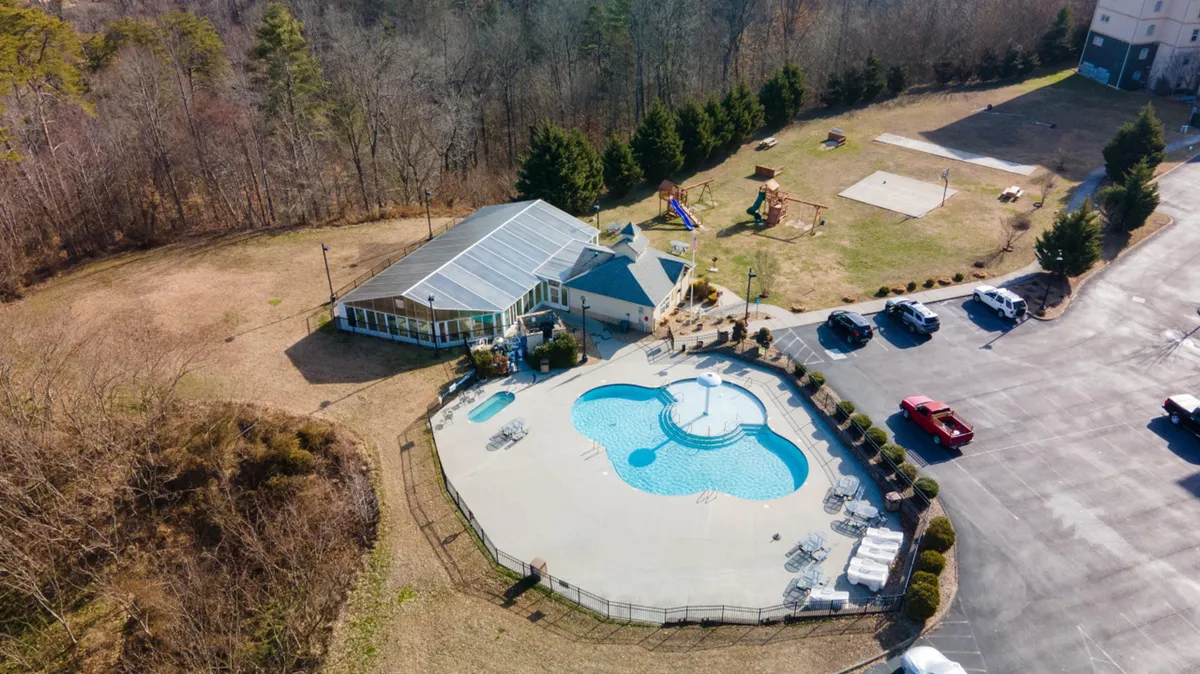 A lively outdoor scene featuring a sparkling swimming pool, nearby playground, and a clubhouse in the background. Children and families gather poolside, with sunbathing and playful activities happening simultaneously, all set in a relaxed, cheerful atmosphere.