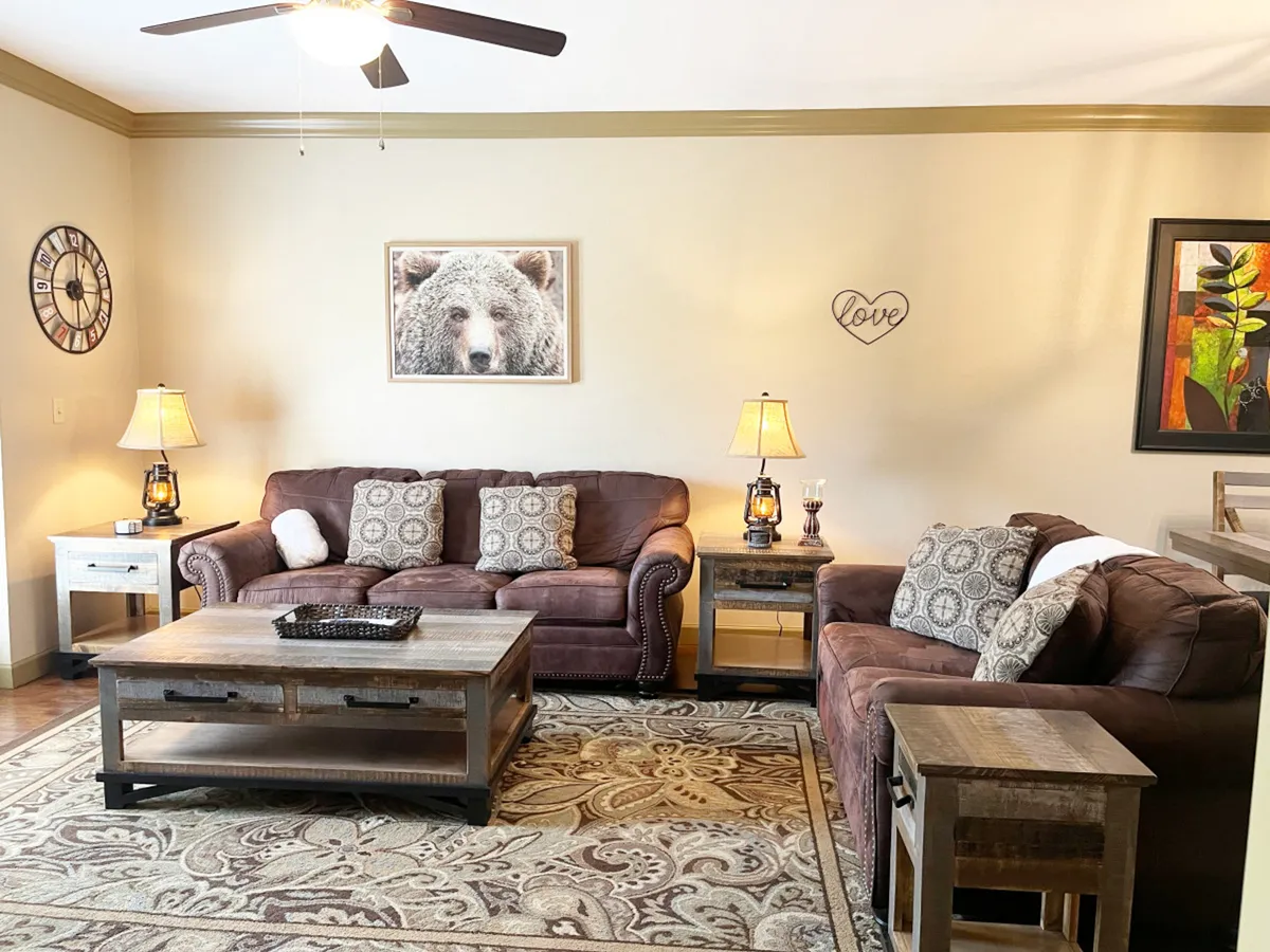A cozy living room featuring leather sofas clustered around a wooden coffee table, with warm lighting and bear-themed artwork on the wall. The space feels inviting and perfect for relaxing, socializing, or enjoying a movie night.