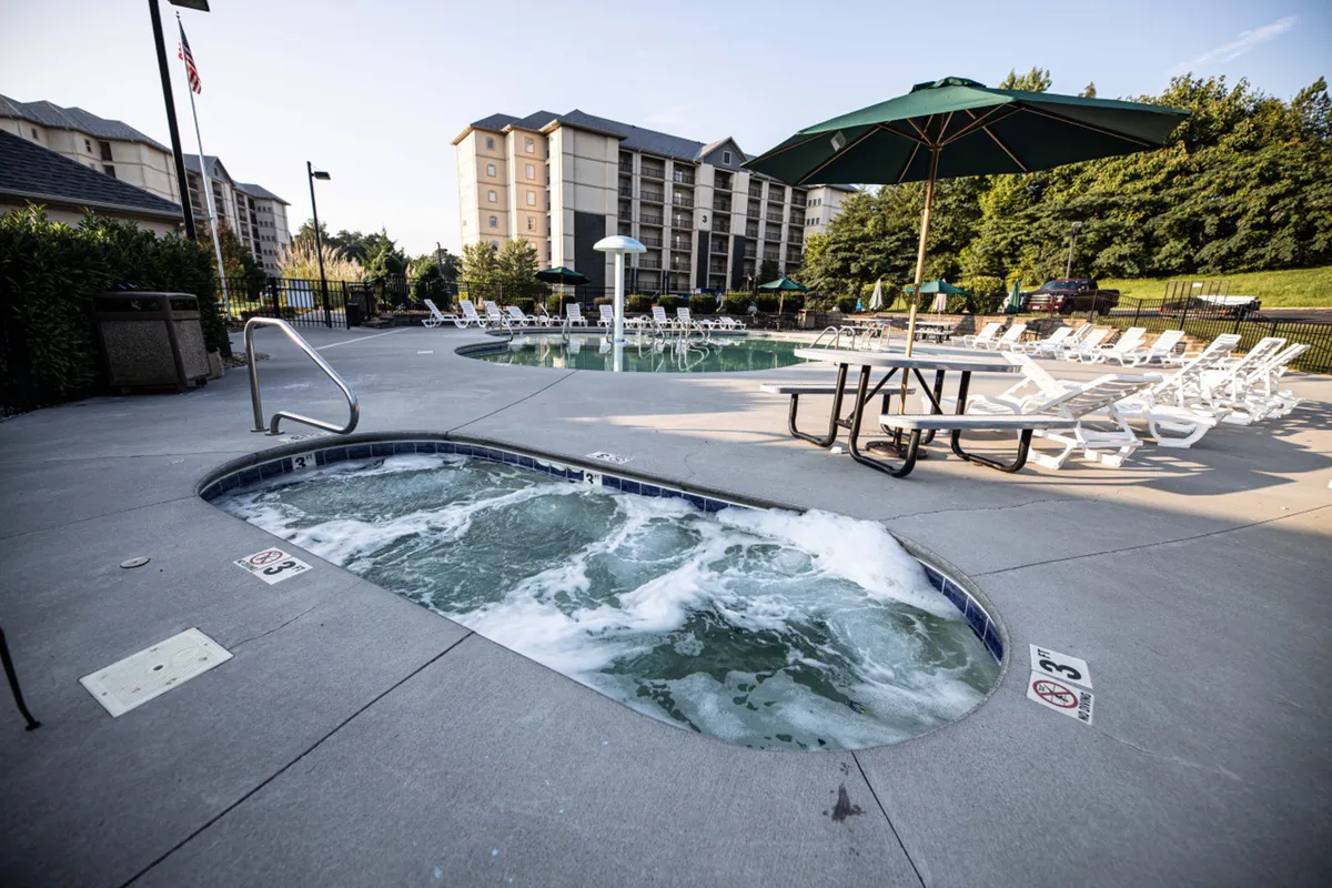 This image features a relaxing outdoor space with a hot tub, swimming pool, lounge chairs, an umbrella, and a picnic table. The area is bathed in natural light and set up for leisure, offering places to soak, swim, sunbathe, or enjoy a meal outside.