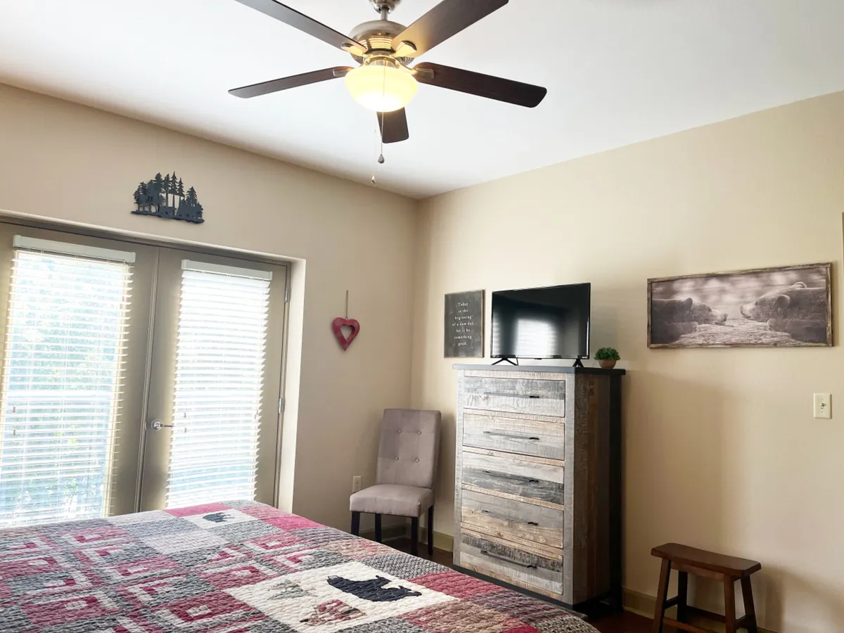 A serene master bedroom with a king-sized bed dressed in a patterned quilt, complemented by a wooden dresser, a ceiling fan, and sliding glass doors that bring in natural light. The space exudes tranquility and comfort.